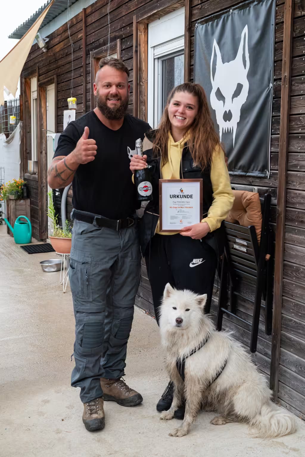 Georg Resch und eine frisch zertifizierte Hundetrainerin mit Urkunde und weißem Hund vor dem Vereinsgebäude